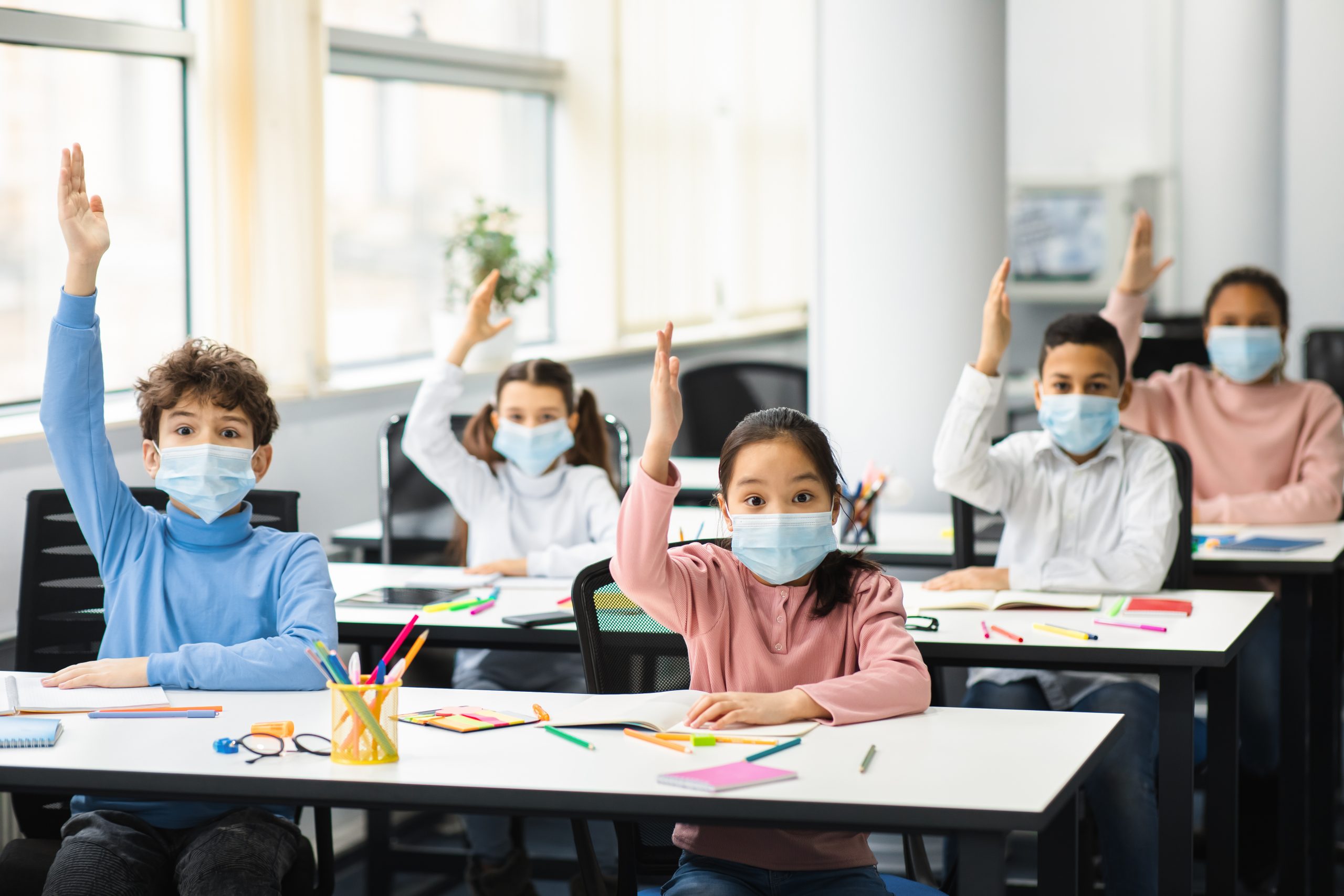 Coronavirus Pandemic And Reopening Schools. Multicultural small school children raising hands at lesson, ready for answer. Diverse pupils wearing disposable surgical masks for protection from virus